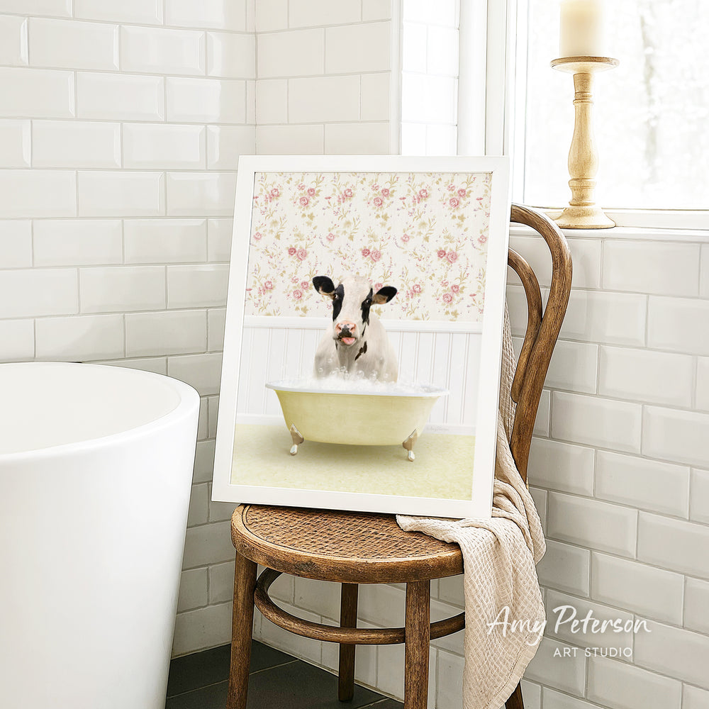 A framed image of a cow sitting in a bathtub, surrounded by a floral pattern, is displayed on a wooden chair in a bathroom setting.