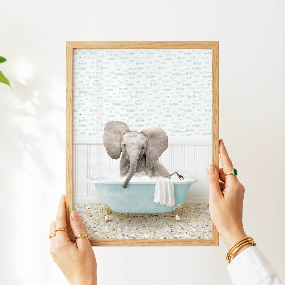A person's hands are holding a framed picture of a baby elephant sitting in a bathtub filled with water, surrounded by fish-themed wallpaper.
