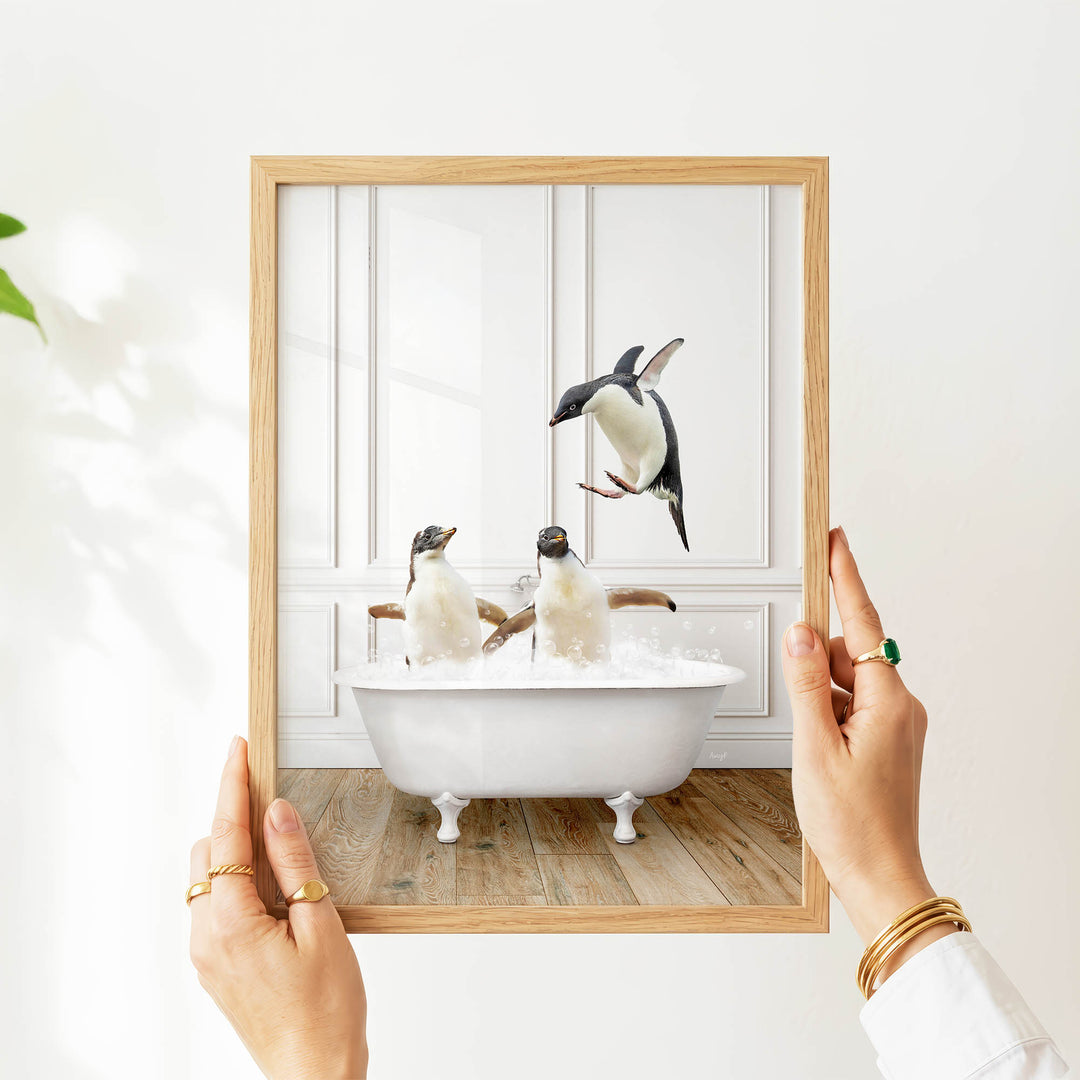 A person's hands are holding a picture frame with a photograph of two penguins sitting in a bathtub, with one penguin flying out of the tub.