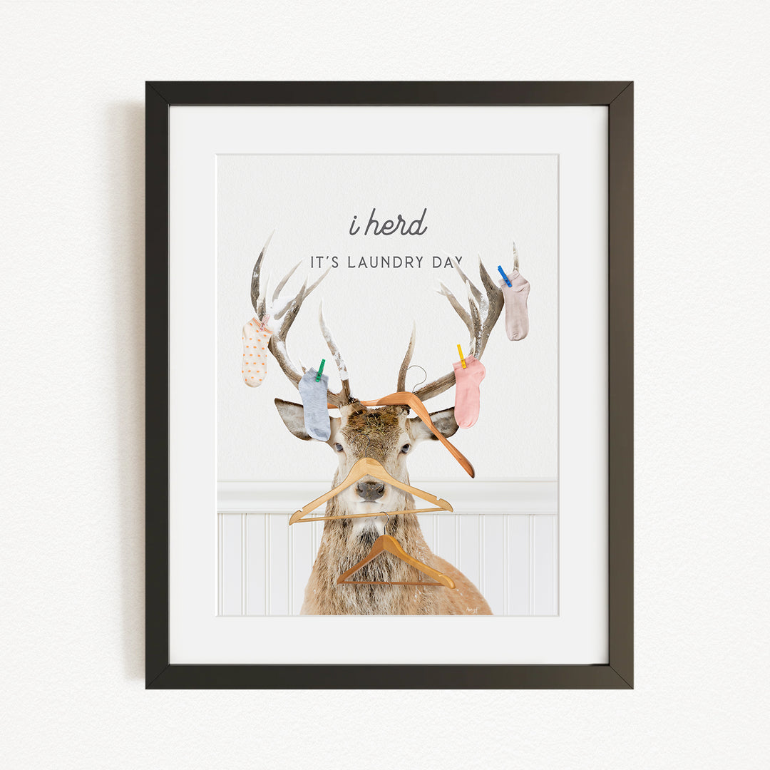 A framed print featuring a deer with antlers holding clothes and hangers, accompanied by the text "It's laundry day".