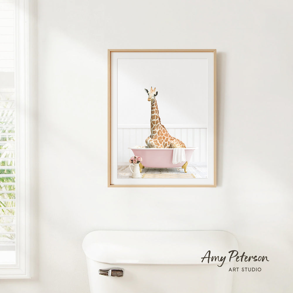 A framed image of a giraffe sitting in a pink bathtub, with a vase of flowers and a white toilet in the foreground.