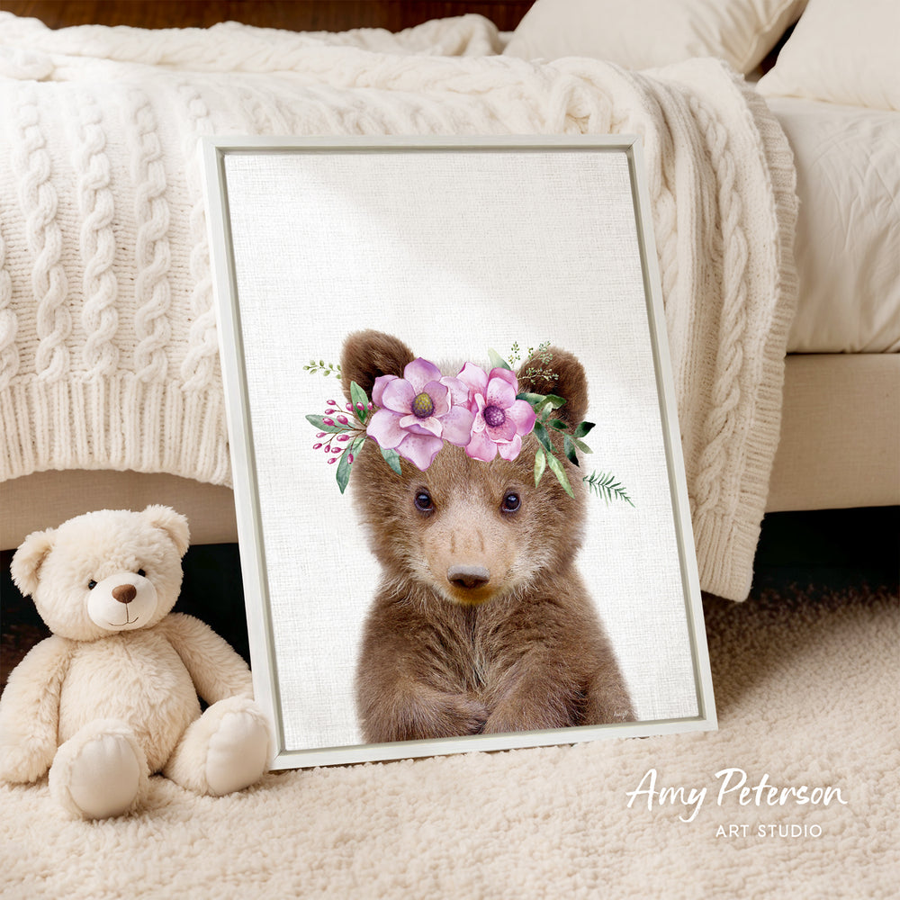 A framed print of a teddy bear wearing a flower crown is displayed on a white bedspread, with a stuffed teddy bear next to it.