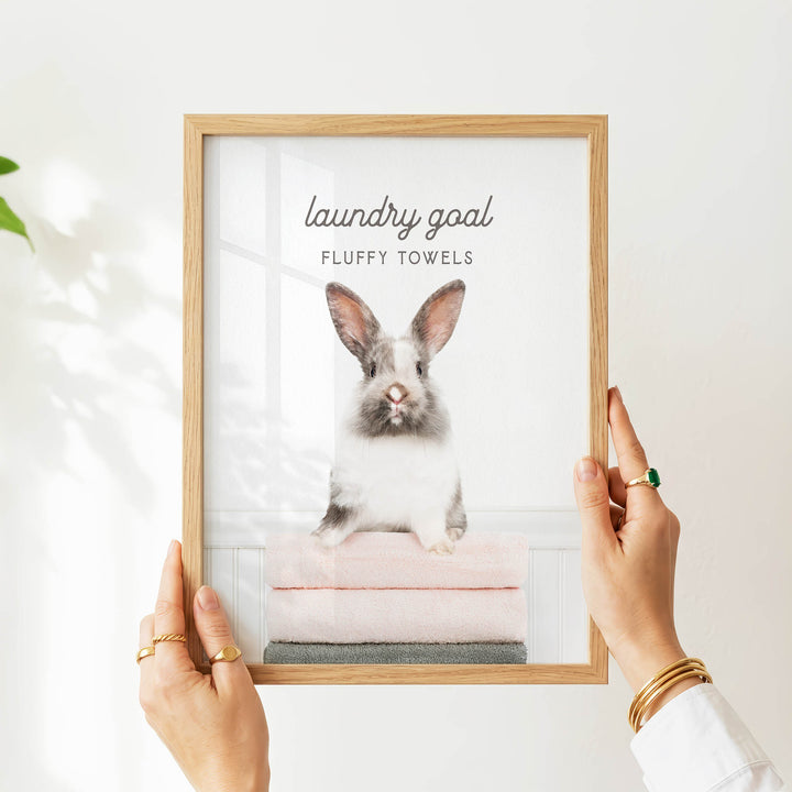 A person is holding a framed picture of a fluffy white rabbit sitting on a stack of pink towels.