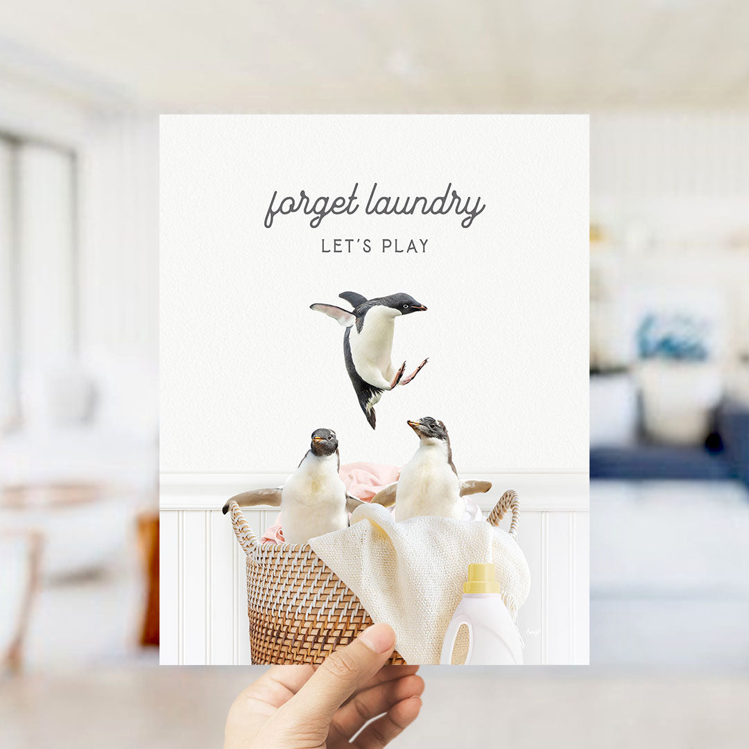 A hand holding a card with a playful penguin image, accompanied by the text "forget laundry, let's play".
