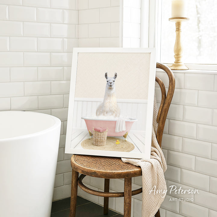 A framed image of a white llama sitting in a pink bathtub, with a basket of bath toys nearby, is displayed on a wooden chair in a bathroom setting.