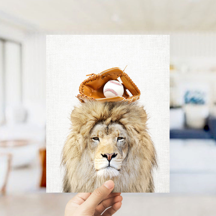 A hand holds up a framed image of a lion wearing a baseball glove and ball on its head, with a blurred background suggesting a living room setting.