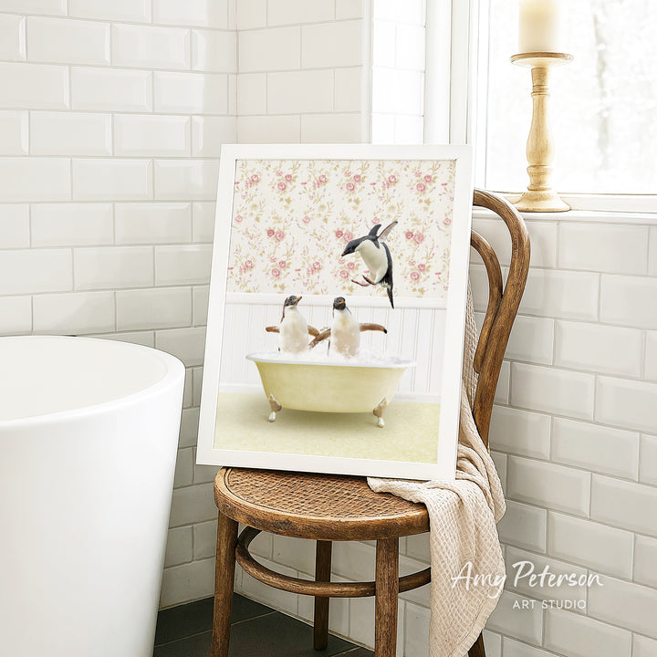 A framed image of two penguins sitting in a bathtub, surrounded by a floral pattern, is displayed on a wooden chair in a bathroom setting.