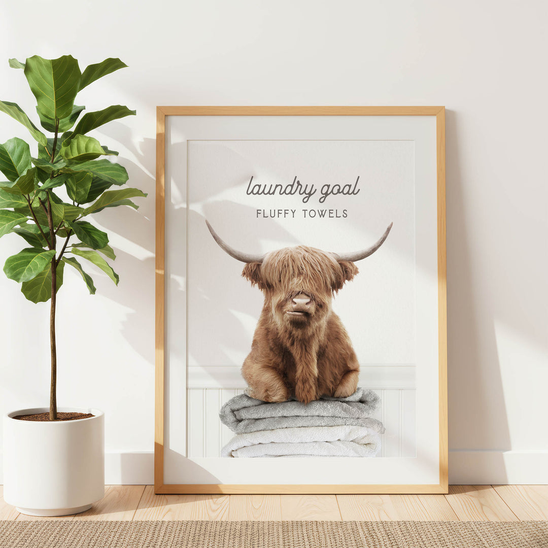 a framed poster of a Highland cow sitting on a stack of fluffy towels, with the text "laundry goal FLUFFY TOWELS" written above it.