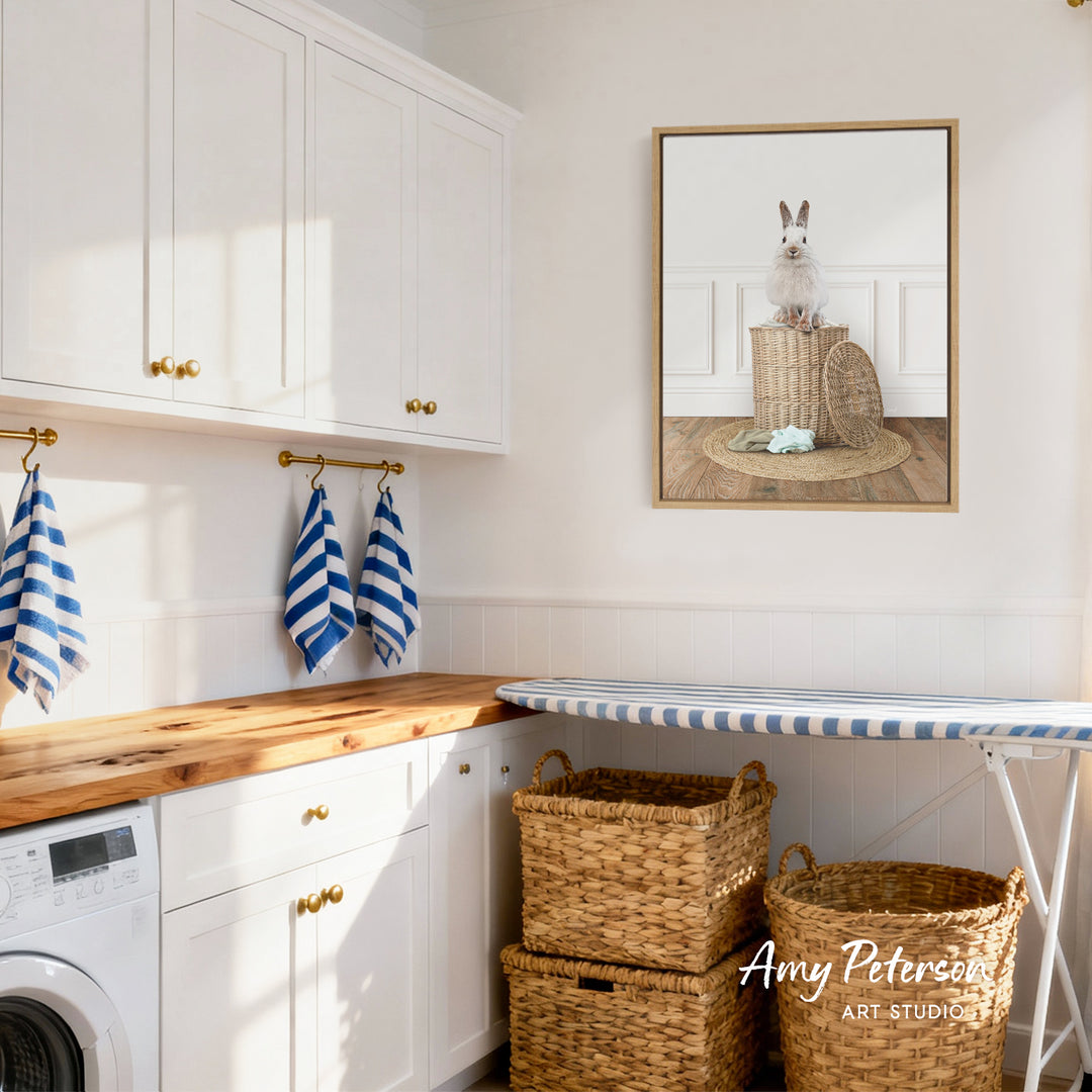 A cozy laundry room with white cabinets, a wooden countertop, and a framed picture of a rabbit on a basket. There are also two woven baskets and a striped ironing board in the room.