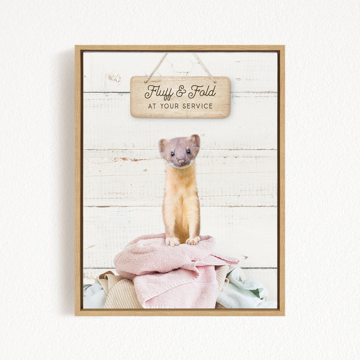 A cute beige cat sitting on a pink blanket, with a wooden sign above that says "Fluffy & Fold at your Service".