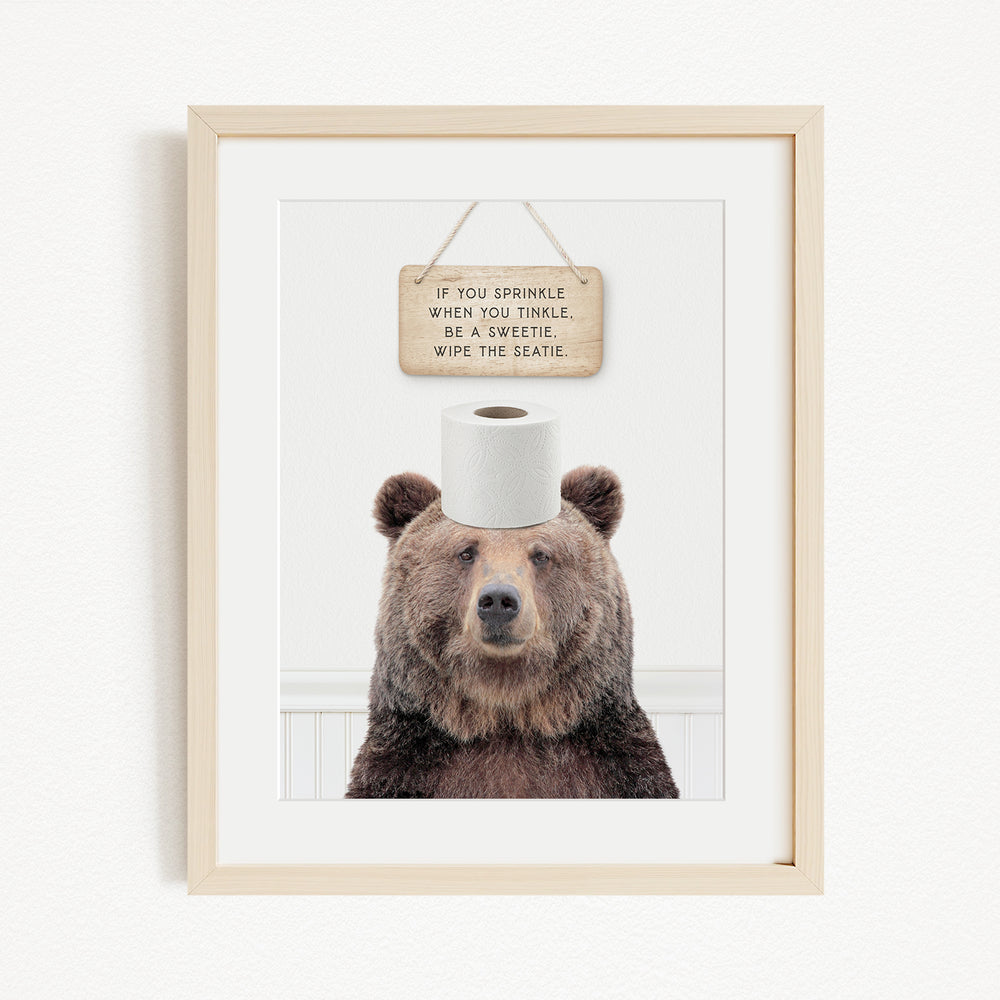 A framed image of a brown bear sitting on a roll of toilet paper, with a wooden sign above it that reads "If you sprinkle when you tinkle, be a twinkle, wipe the seattle."