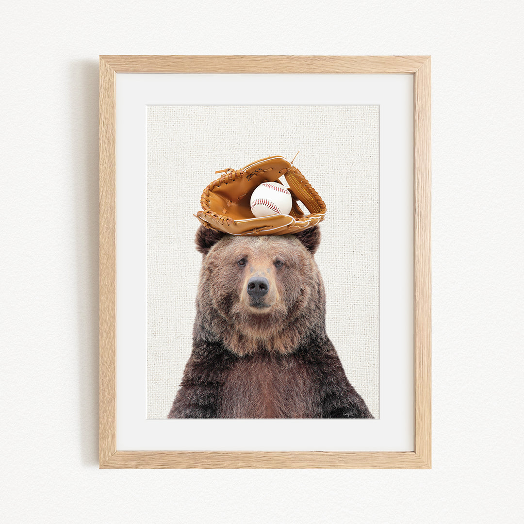 A framed image of a brown bear wearing a baseball glove on its head, with a baseball resting on top of the glove.