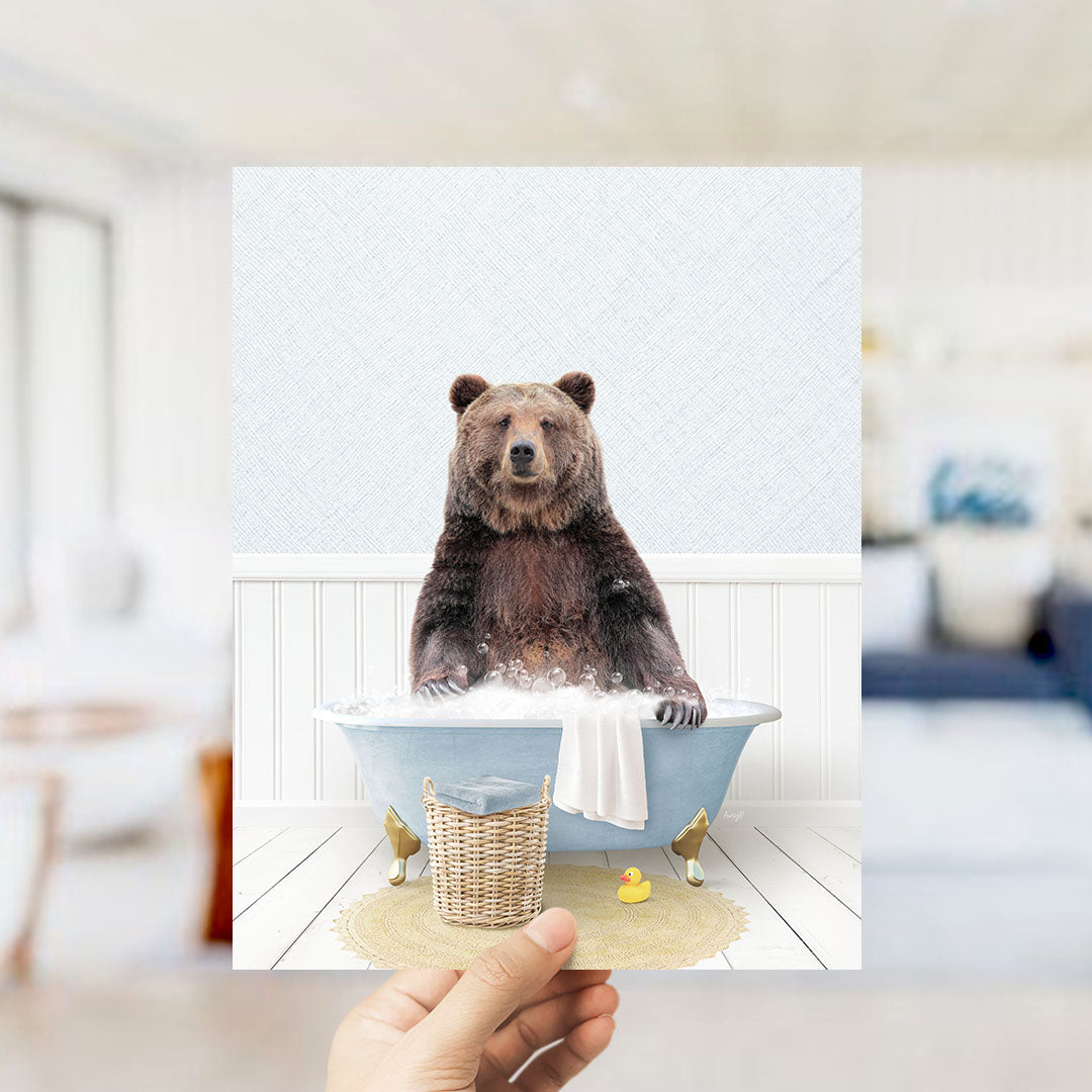 A hand holds up a framed image of a brown bear sitting in a bathtub filled with bubbles, surrounded by a yellow rubber duck and a wicker basket.