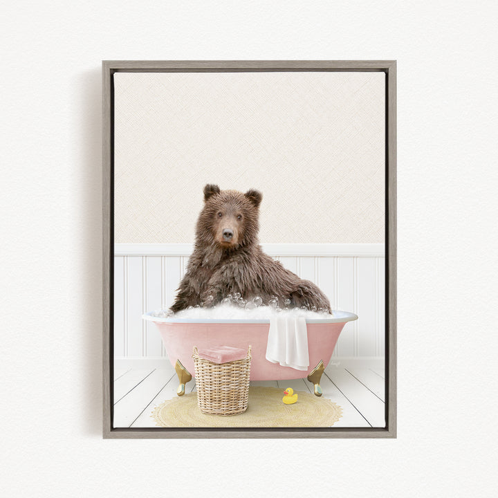 A brown bear sitting in a pink bathtub filled with bubbles, surrounded by a yellow rubber duck and a wicker basket.