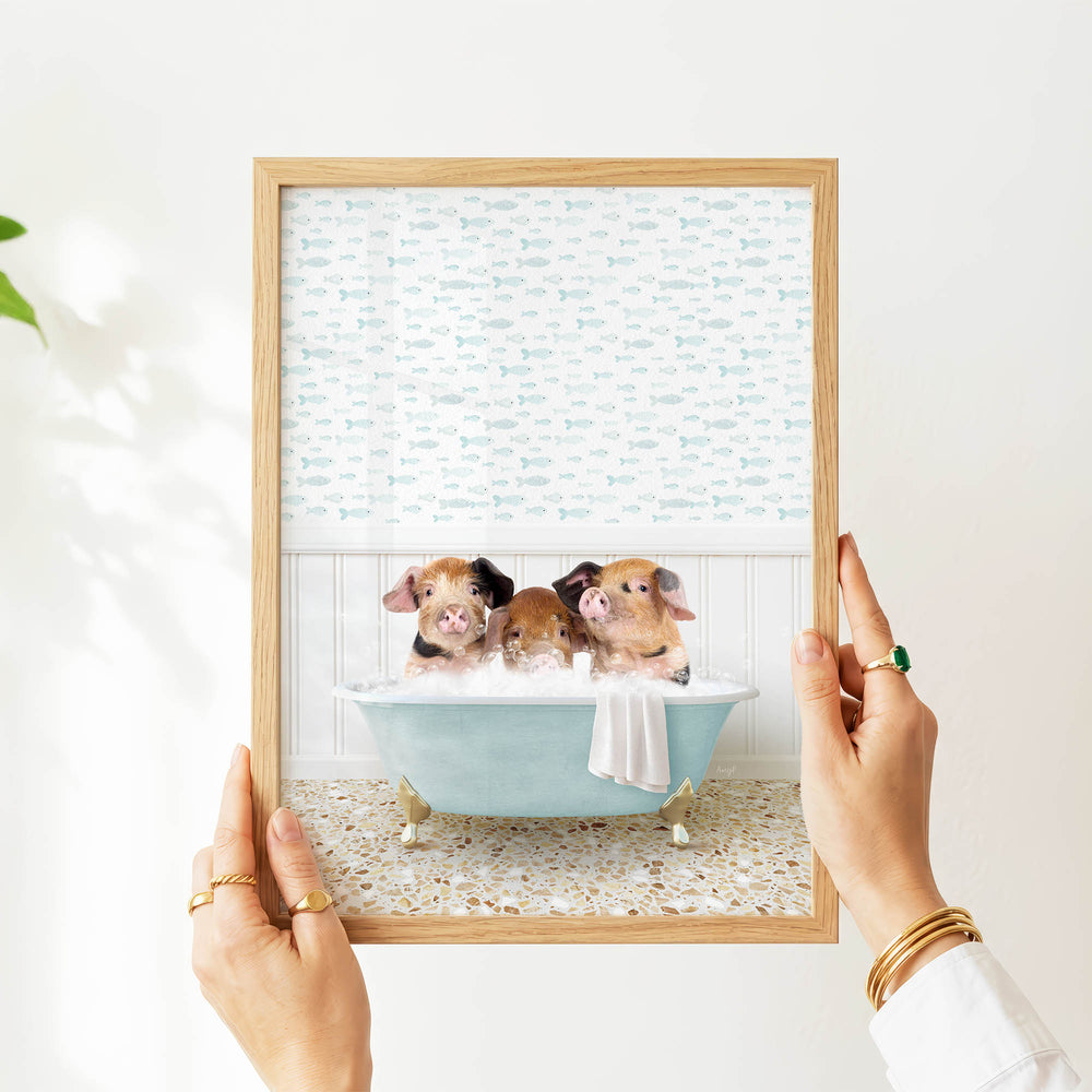 A person's hands are holding a framed picture of three adorable pigs sitting in a bathtub filled with bubbles, surrounded by a pattern of fish.