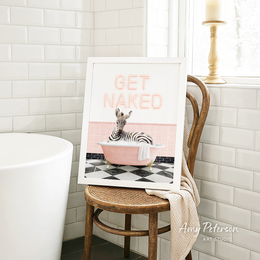 A framed print of a zebra sitting in a pink bathtub with the text "GET NAKED" on it, placed on a wooden chair in a bathroom setting.