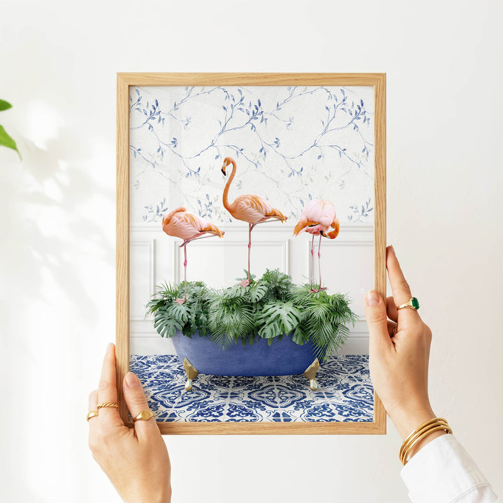 A person's hands are holding a framed picture of three pink flamingos standing in a blue bathtub filled with green plants.