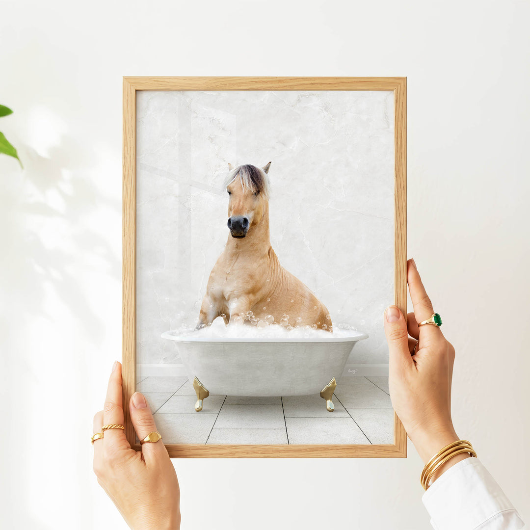 A person's hands are holding a framed picture of a horse sitting in a bathtub filled with bubbles.
