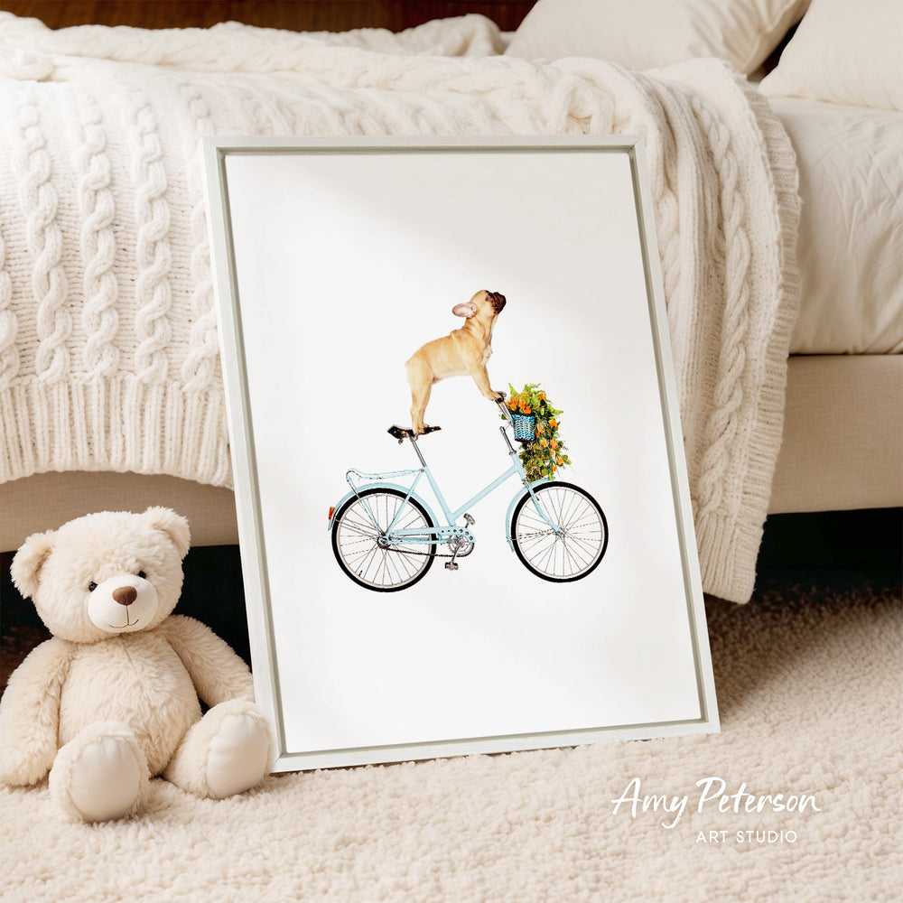 A framed illustration of a dog riding a bicycle, with a basket of flowers attached to the back. The dog is sitting on a blue bicycle with a basket on the front. Next to the framed illustration is a stuffed teddy bear.