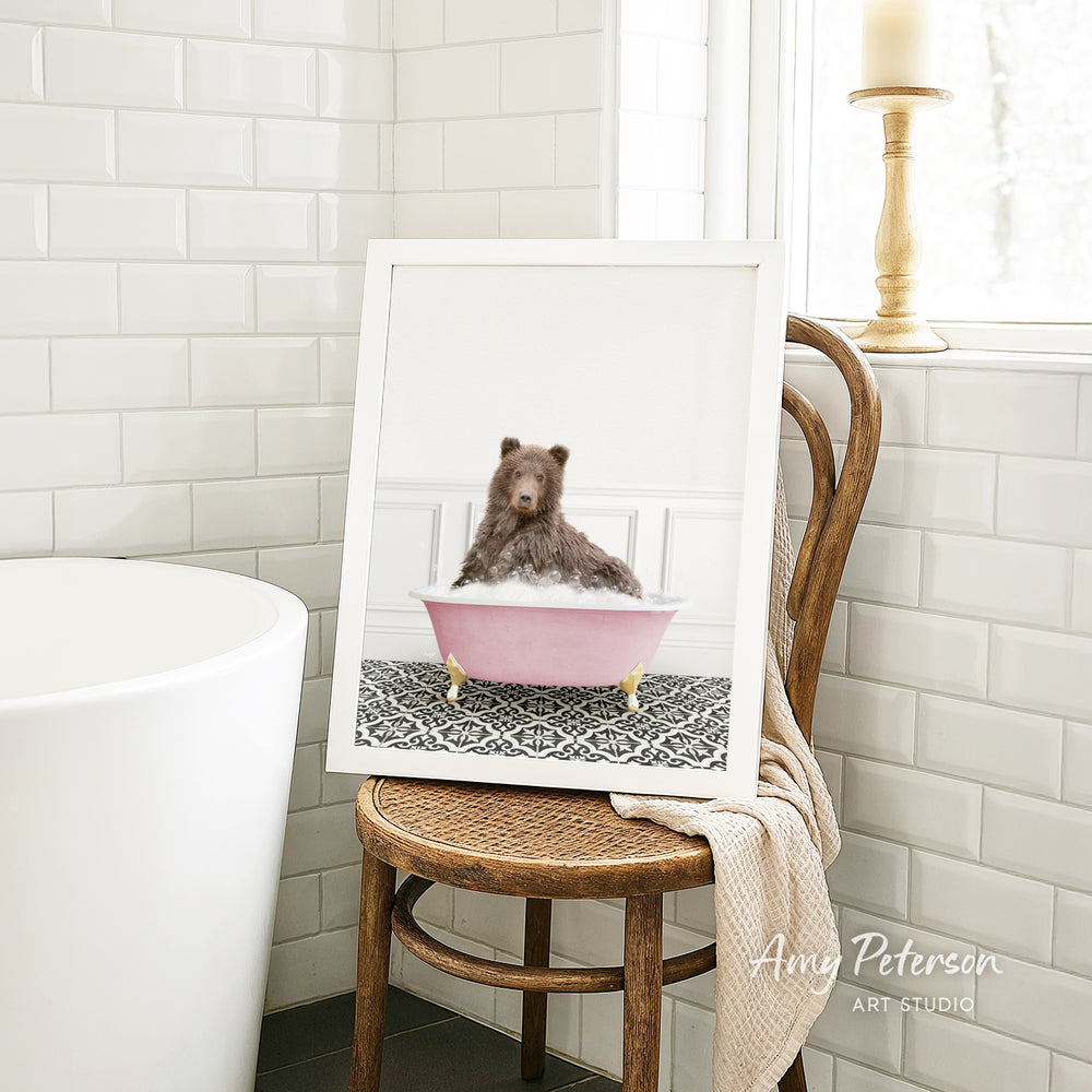 A framed image of a brown bear sitting in a pink bathtub, with a towel draped over the back of a wooden chair in a bathroom setting.