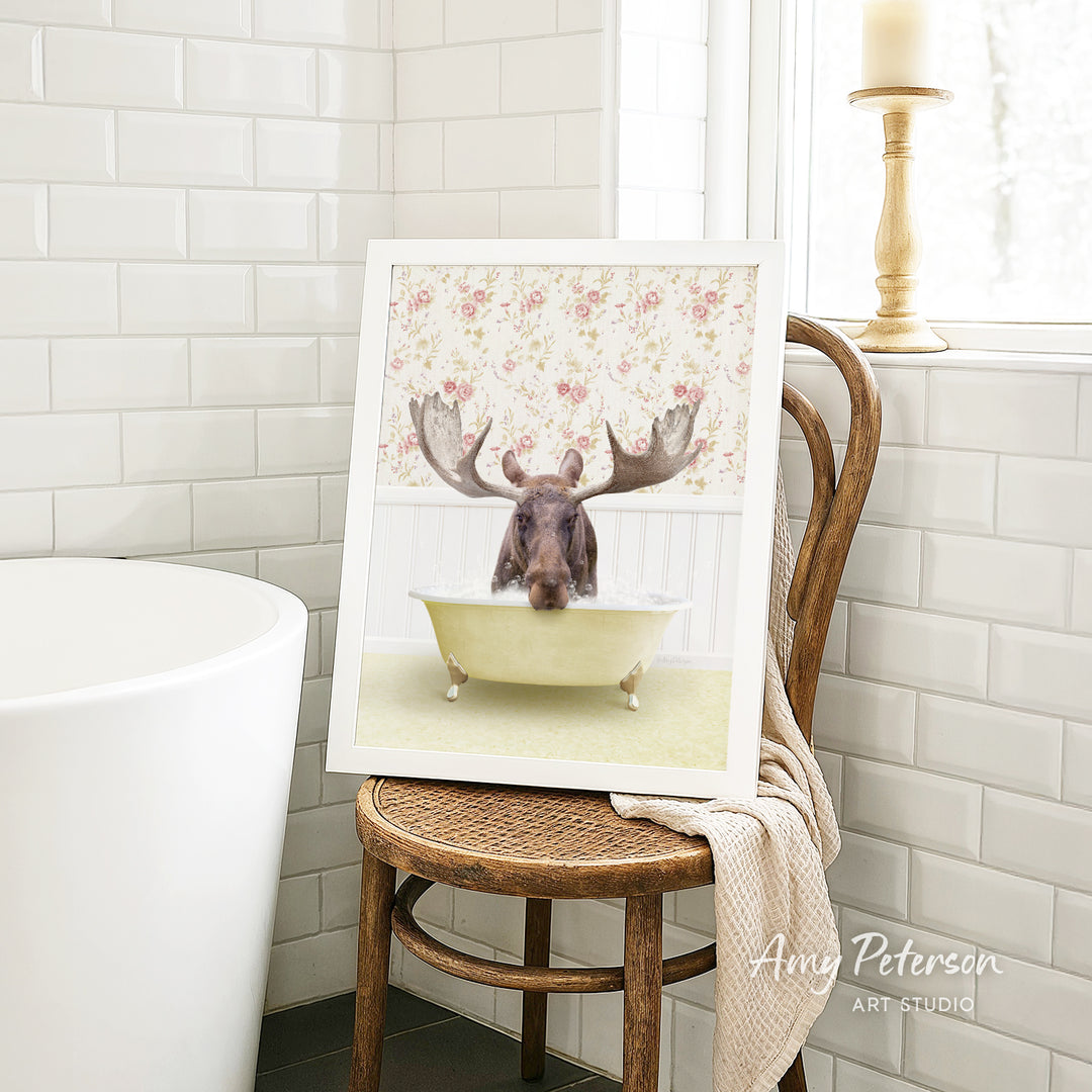 A framed image of a moose sitting in a bathtub, with its antlers raised, is displayed on a wooden chair in a bathroom setting.