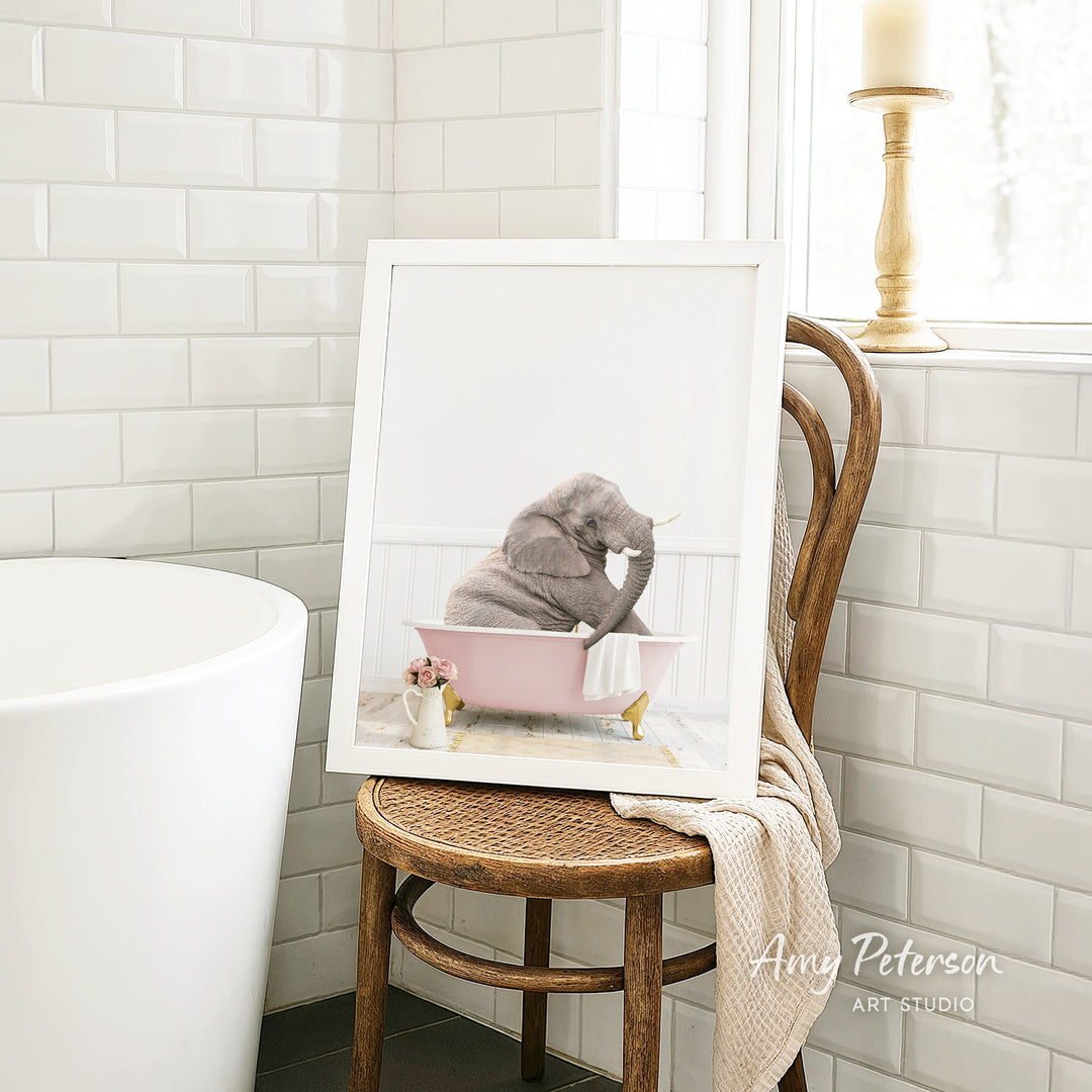 A framed photograph of a gray elephant sitting in a pink bathtub, with a white towel and flowers nearby, is displayed on a wooden chair in a bathroom setting.