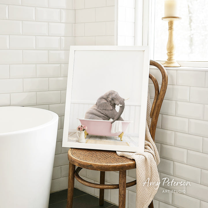 A framed photograph of a gray elephant sitting in a pink bathtub, with a white towel and flowers nearby, is displayed on a wooden chair in a bathroom setting.