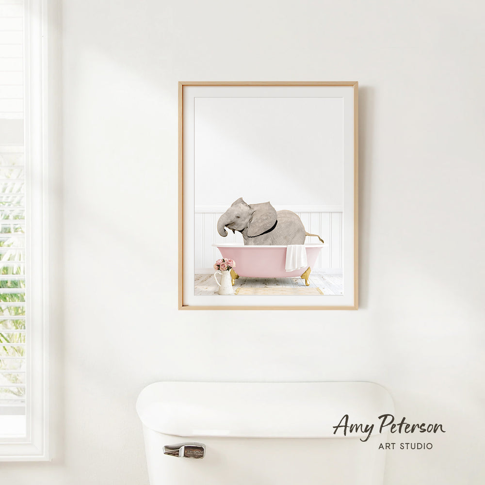 A framed image of an elephant sitting in a pink bathtub, with a vase of flowers and a white toilet in the foreground.