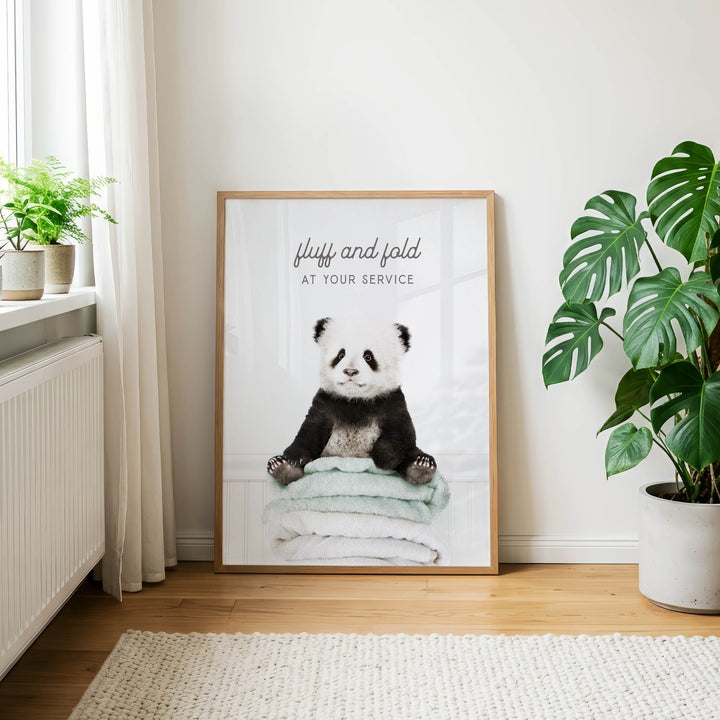 A framed poster of a panda sitting on a stack of towels is displayed on a wall in a room with a window, plants, and a rug.