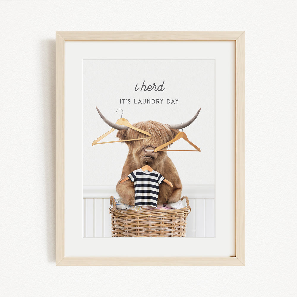 A framed image depicting a Highland cow sitting in a wicker basket, surrounded by clothes and a sign that says "It's laundry day".