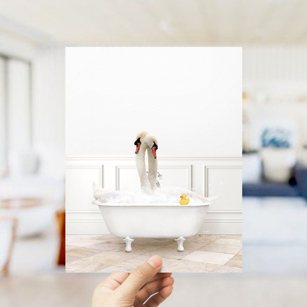 A hand holds up a framed image of a white bathtub with two swans sitting in it, one of which is holding a yellow rubber duck.