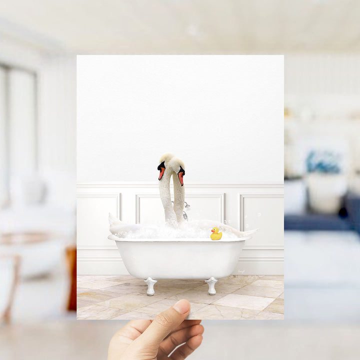 A hand holds up a framed image of a white bathtub with two swans sitting in it, one of which is holding a yellow rubber duck.