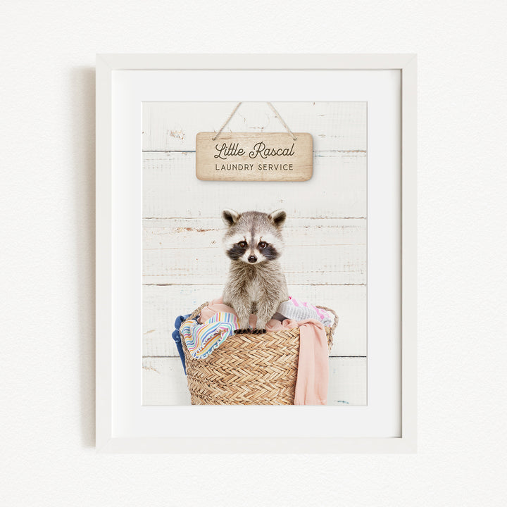 A raccoon sitting in a woven laundry basket, with a sign above that reads "Little Racal Laundry Service".
