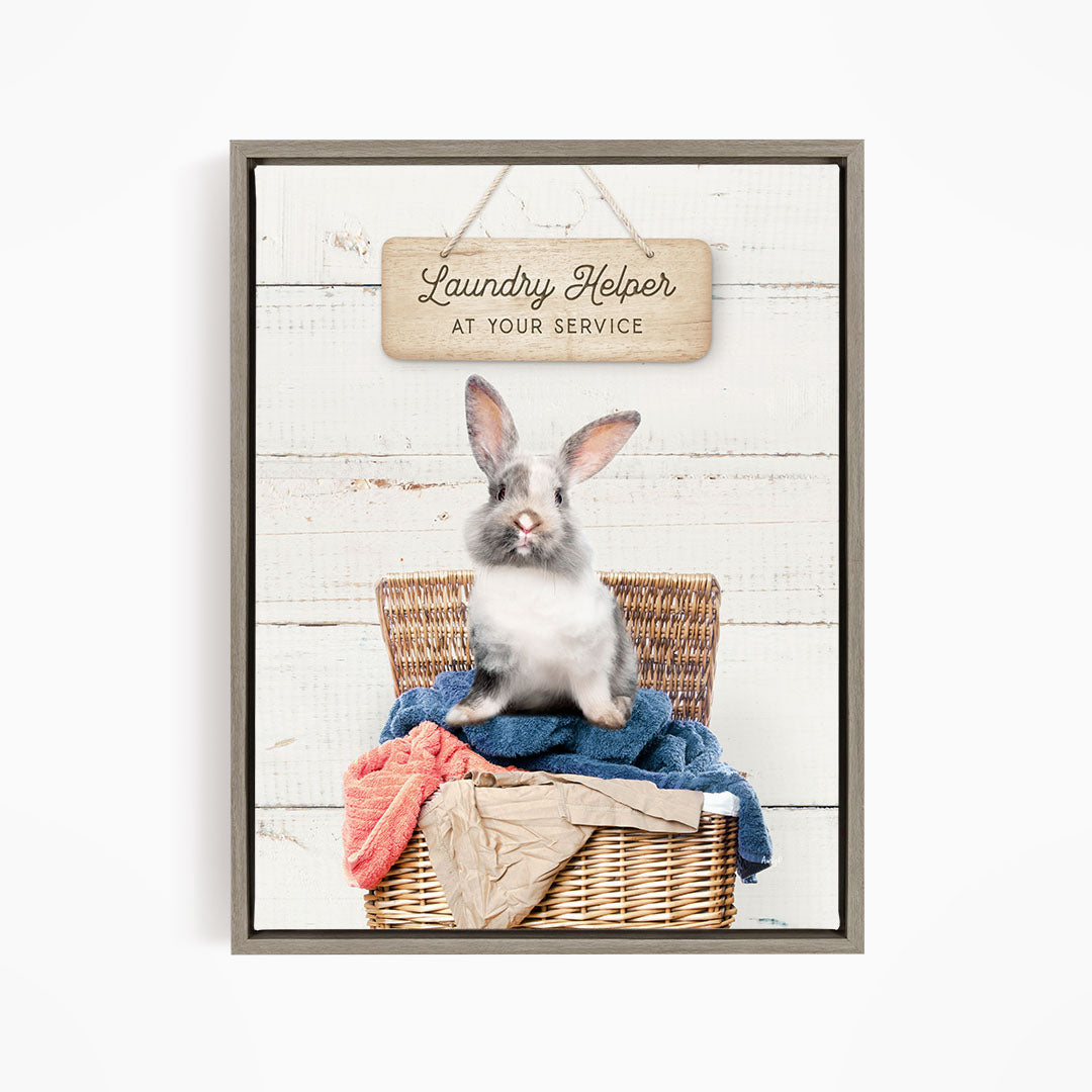A gray and white rabbit sitting in a wicker laundry basket, with a wooden sign above that reads "Laundry Helper at your service".