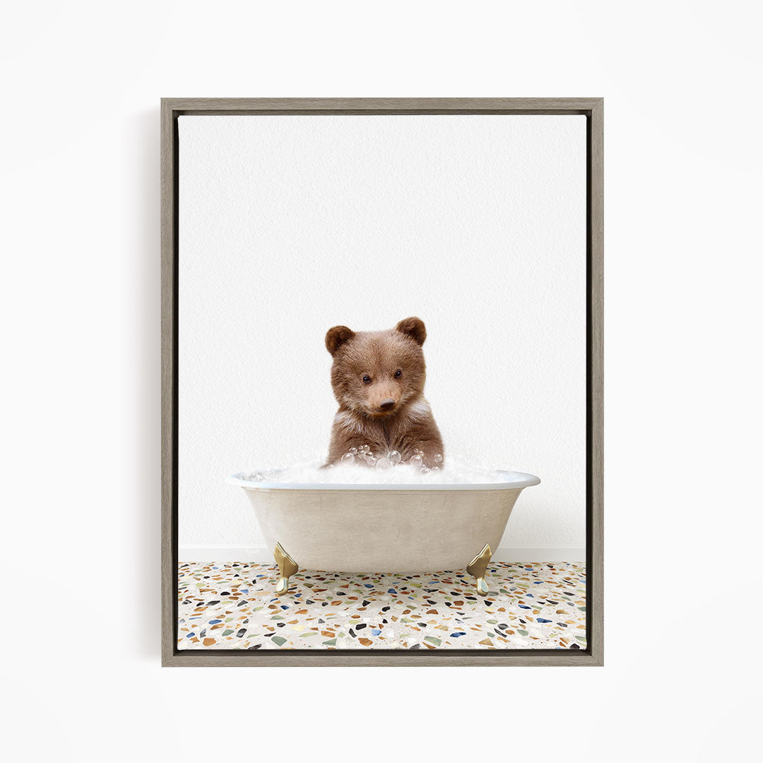 A cute brown bear cub sitting in a white bathtub filled with water, looking directly at the camera.
