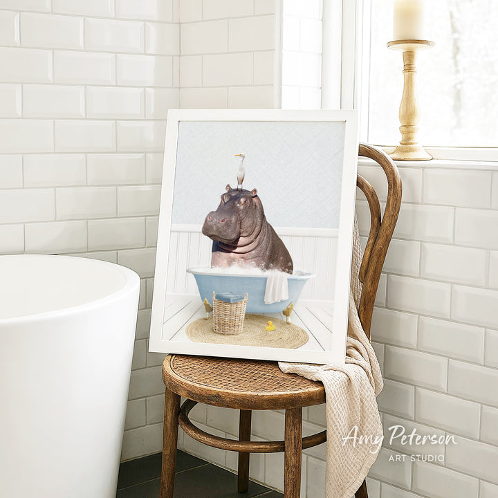 A framed image of a hippopotamus sitting in a bathtub, surrounded by a towel and a basket of bath toys, is displayed on a wooden chair in a bathroom setting.