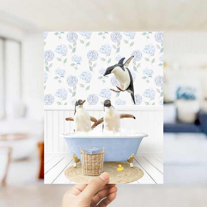 A hand holds up a framed image depicting two penguins sitting in a bathtub filled with water, surrounded by a blue and white floral pattern.