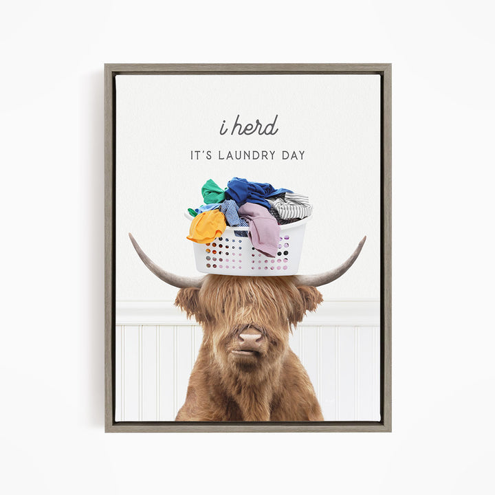 A brown cow with long hair is sitting on a white laundry basket, which is filled with clothes. The cow has a basket on its head, and there is text above the image that says "i herd IT'S Laundry Day".