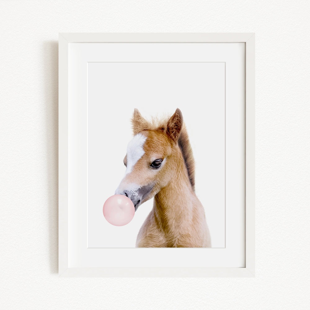 A framed image of a brown horse blowing a bubble with a pink bubble gum bubble.
