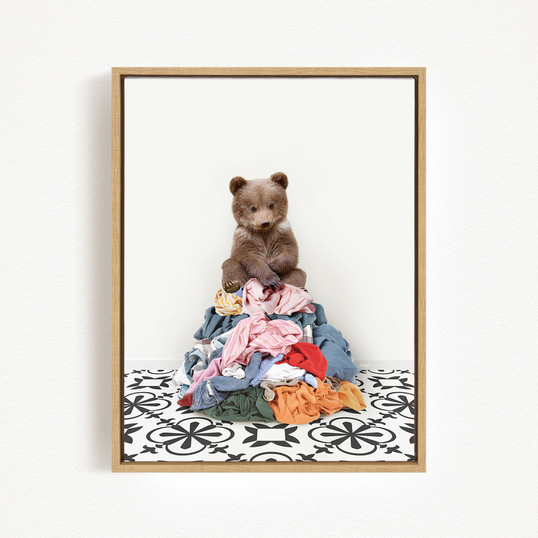 A brown bear sits atop a pile of clothes on a patterned floor.