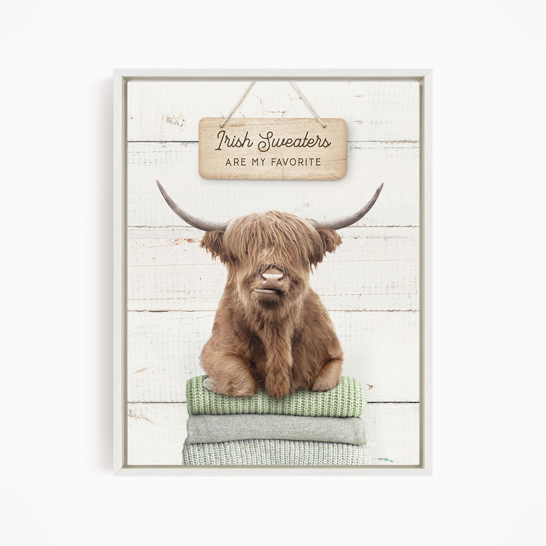 A brown Highland cow with large horns is sitting on a stack of green and gray blankets, with a sign above that says "Irish Sweaters are my favorite".