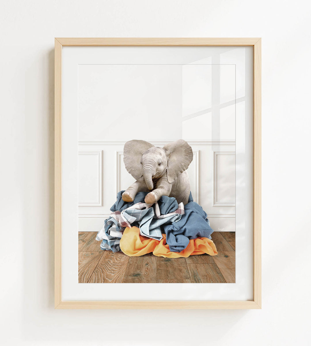 A framed image of a stuffed elephant sitting on a pile of clothes on a wooden floor.