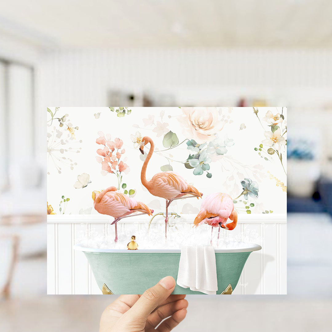A hand is holding up a framed image of three pink flamingos standing in a bathtub filled with water and bubbles, surrounded by a floral pattern.