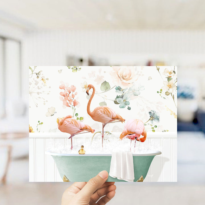 A hand is holding up a framed image of three pink flamingos standing in a bathtub filled with water and bubbles, surrounded by a floral pattern.