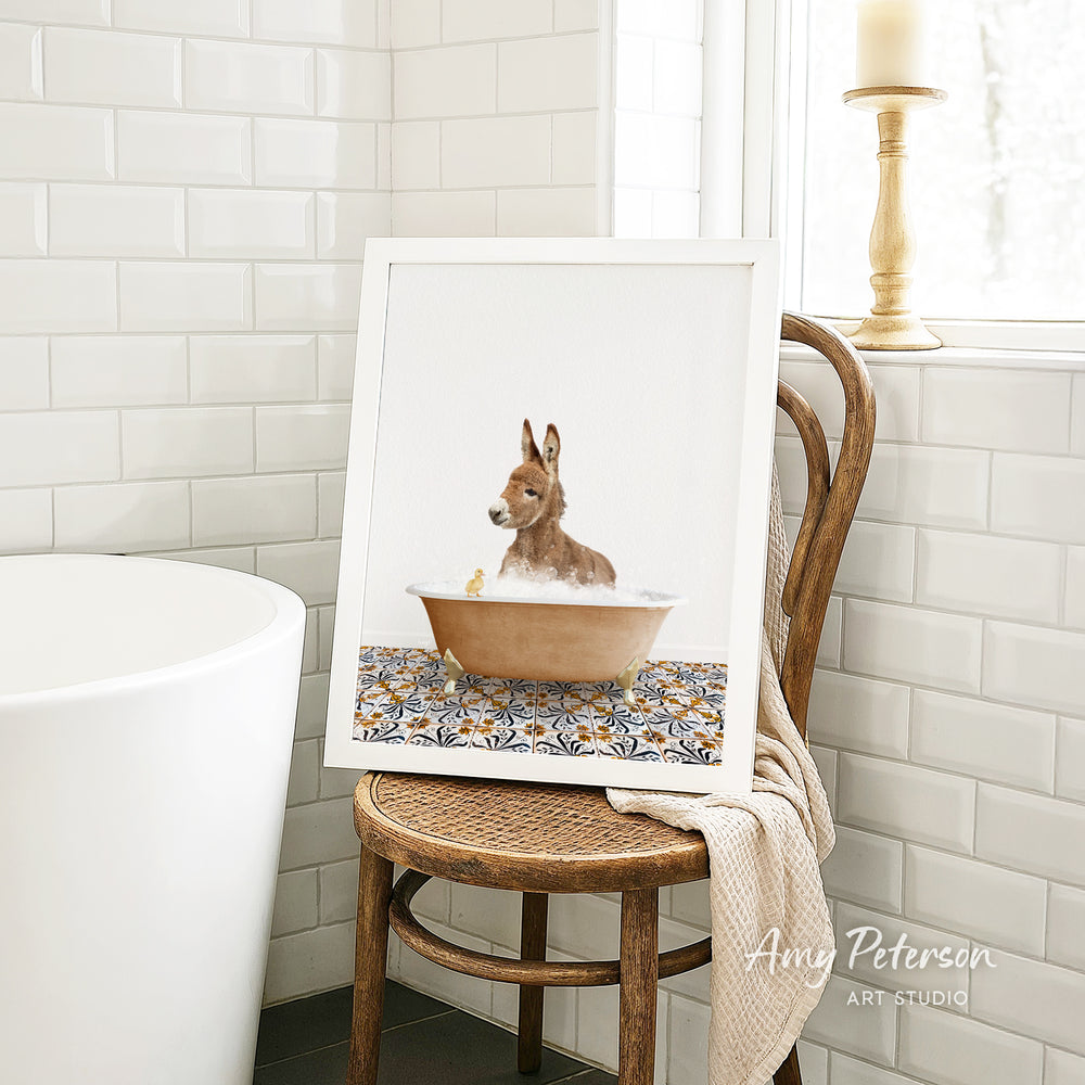 A framed print of a brown rabbit sitting in a bathtub is displayed on a wooden chair in a bathroom setting.