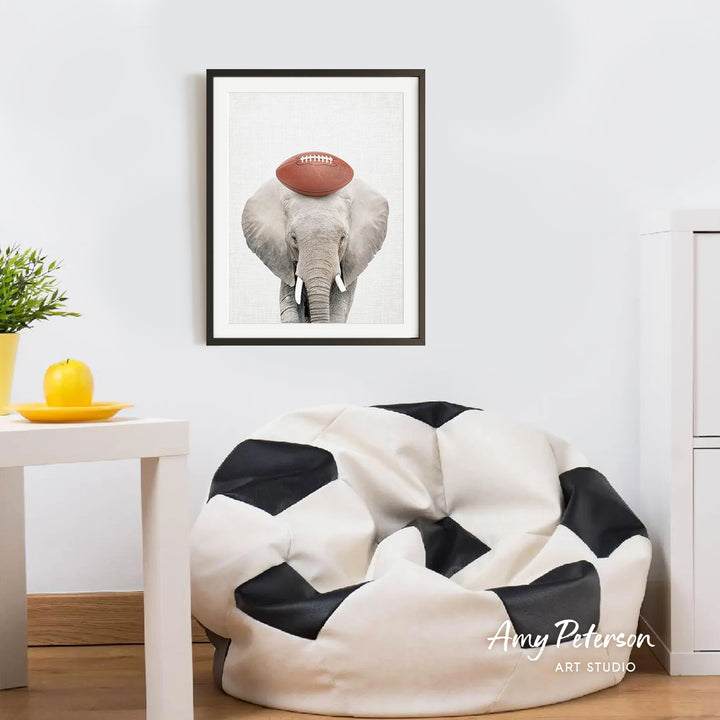 a framed picture of an elephant with a football on its head, hanging on a white wall above a black and white bean bag chair.
