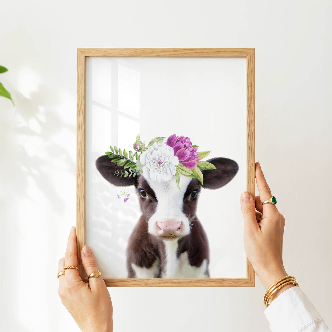 A person's hands are holding a wooden frame with a picture of a cow wearing a flower crown.