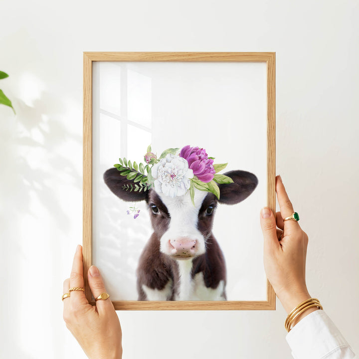 A person's hands are holding a wooden frame with a picture of a cow wearing a flower crown.