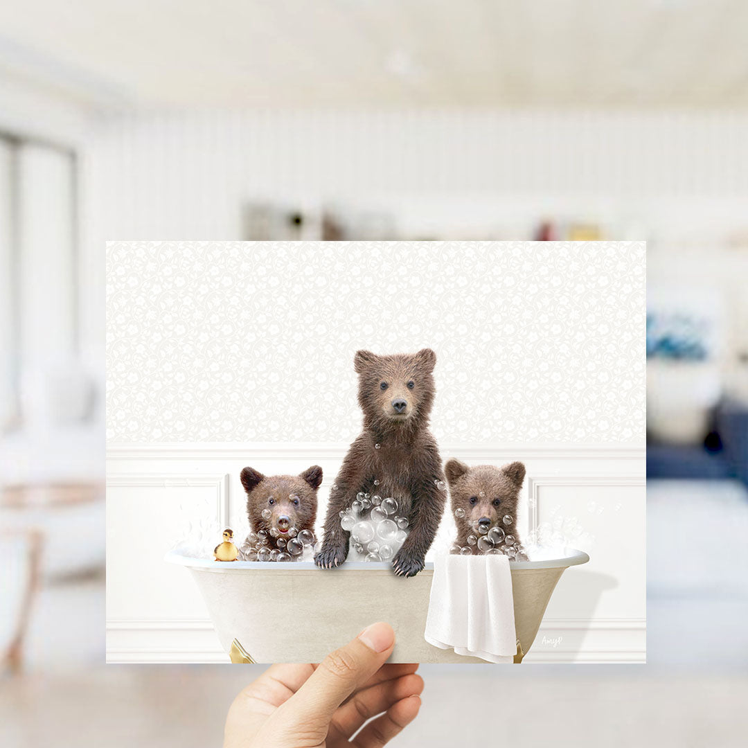 A hand holds up a framed image depicting three adorable brown bears sitting in a bathtub filled with bubbles, creating a whimsical and playful scene.