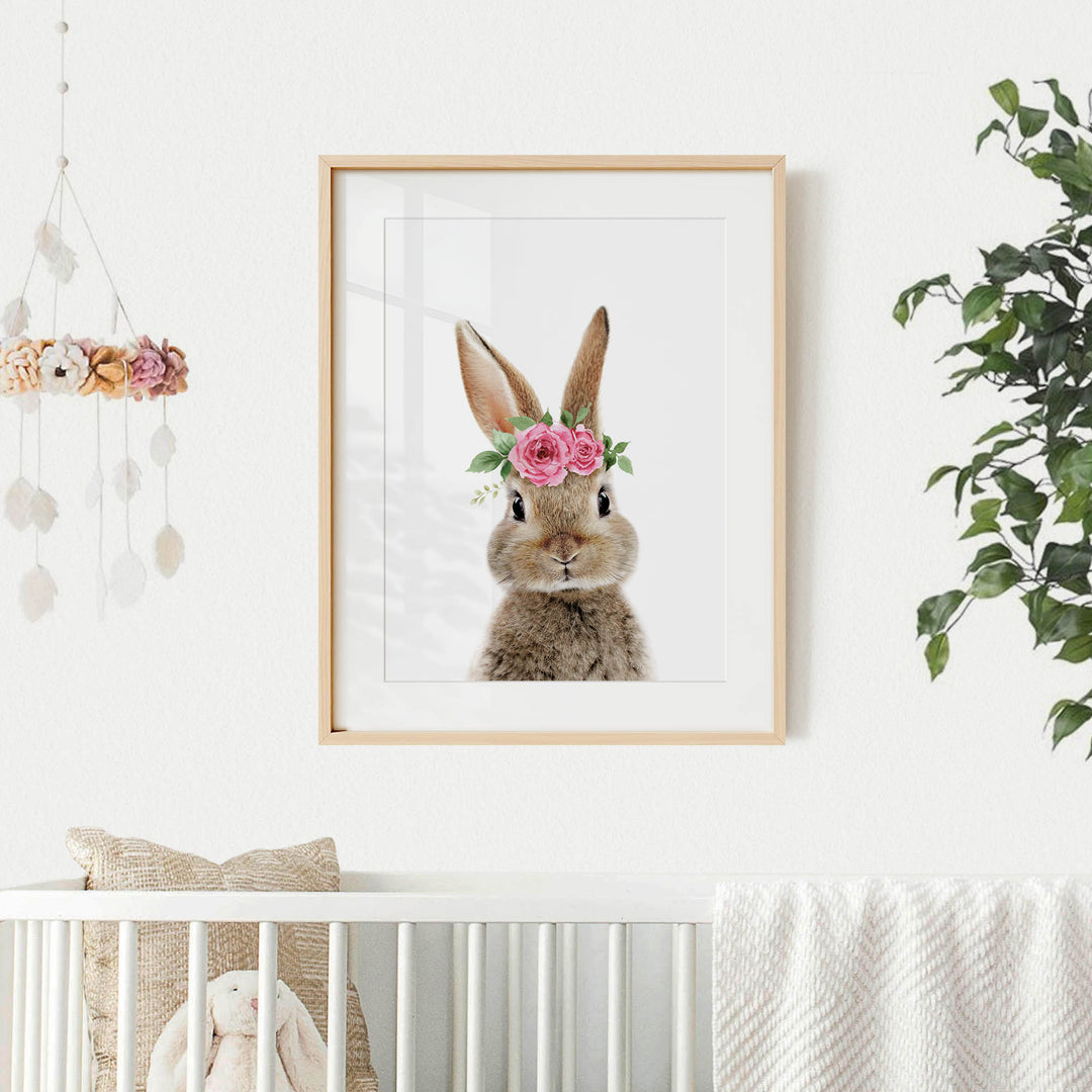 A framed picture of a bunny wearing a flower crown is hanging on a white wall above a white crib.